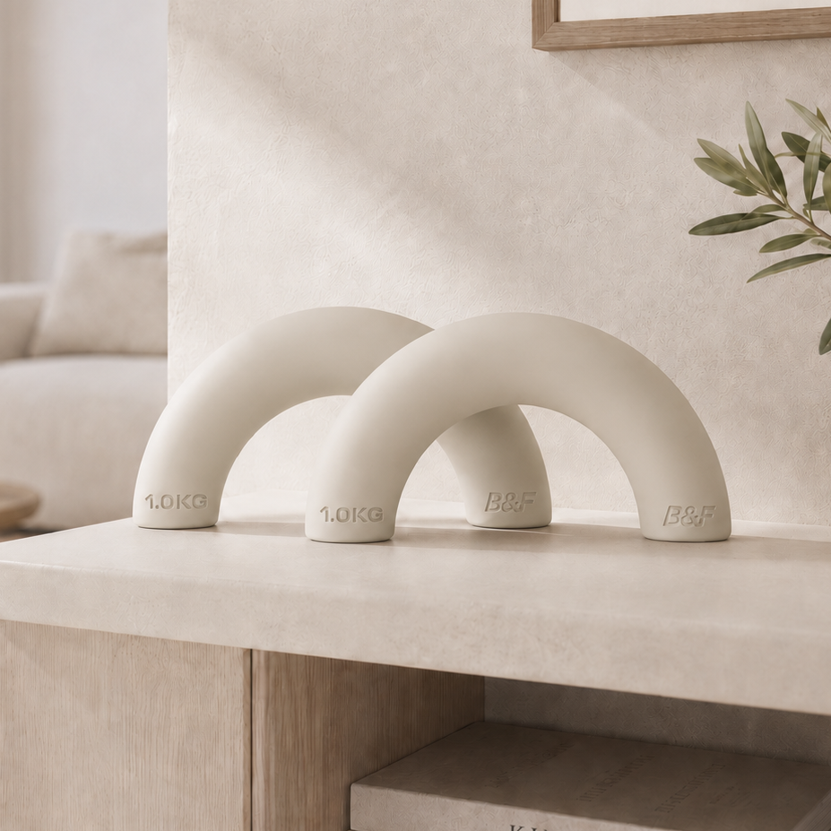 Two matte off-white arched decorative objects displayed on a light stone console table in a softly styled interior room. The scene includes framed wall art, a sofa, plants, and books, with no people or animals present.